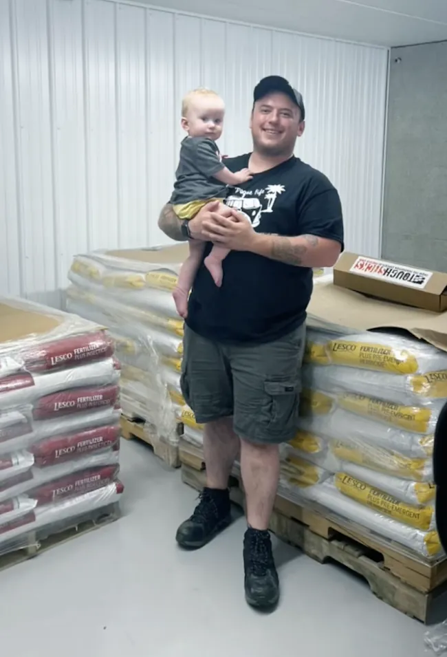 Andrew Duval holding his daughter Elora next to pallets of lawn products